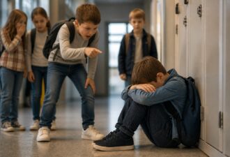 Шкільний коридор: одна дитина сидить біля шафок, закривши обличчя, поруч одноліток агресивно вказує на неї, інші діти спостерігають. Напружена атмосфера, фокус на емоціях і взаємодії, м’яке природне світло без зайвих деталей.